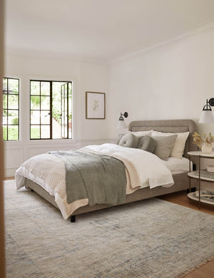 The Ismenia distressed slate-toned persian rug with fringed ends lays in a bedroom with a gray-framed bed, white accented walls, and sage-toned throw pillows and blankets