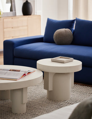 The Anja indoor and outdoor round side table sits atop a grid-patterned rug in front of a deep blue velvet sofa