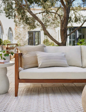 Marchesa khaki indoor and outdoor pillow with tasseled corners sits on a natural linen sofa in an outdoor space with a woven cream rug