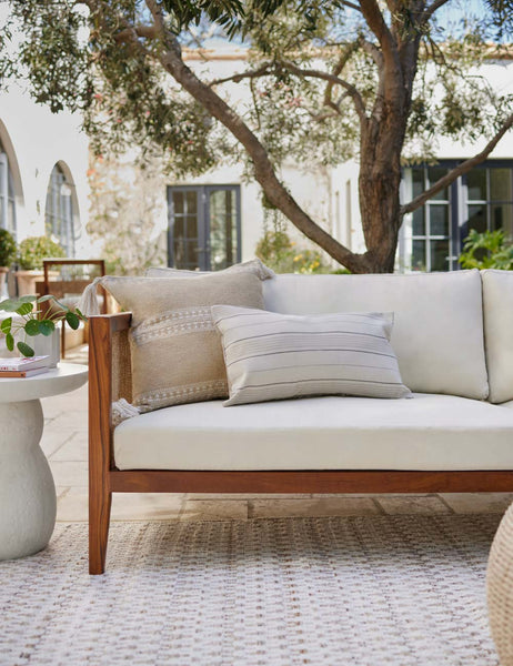 #color::khaki #style::square | Marchesa khaki indoor and outdoor pillow with tasseled corners sits on a natural linen sofa in an outdoor space with a woven cream rug