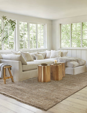 The Kenzi sand rug lays in a bright living room under two plus shaped ottomans, a sectional sofa, and a wooden stool