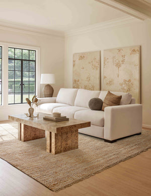 The Kenzi sand rug lays in a living room under a burl wood coffee table and ivory linen sofa near two large wall arts