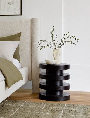 The Pentwater Four-Tiered Black Round Side Table sits next to a natural linen-framed bed and an olive plush rug