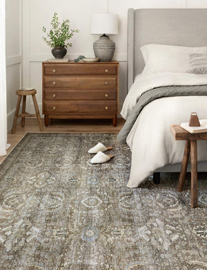 Amalia distressed traditional rug lays under a gray linen framed bed in a bedroom with a wooden bedside dresser, stool, and bench