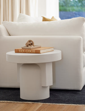 The Anja indoor and outdoor round side table sits next to a white linen couch with a books and sculptural decor atop it