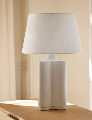 The Duffy white table lamp sits on a wooden side table in a room with warm lighting
