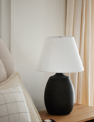 The Pratt black table lamp sits on a wooden nightstand next to a linen framed bed with patterned throw pillows