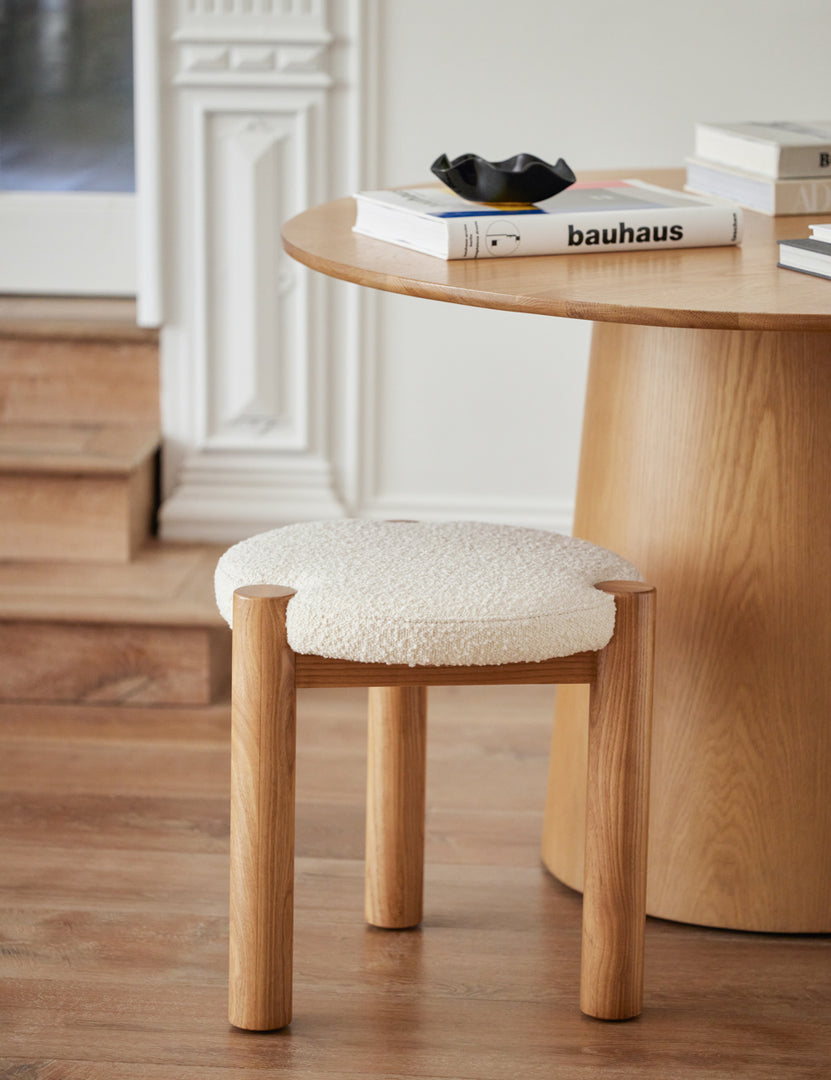 | Kittredge compact stool sits in a room underneath a circular wooden table with a ribbed bowl and stack of books