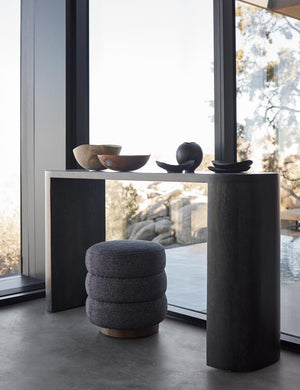 The Luna black mango wood oval console table sits in front of floor to ceiling windows with hand-carved wooden bowls sitting atop it and a gray fabric stool underneath.