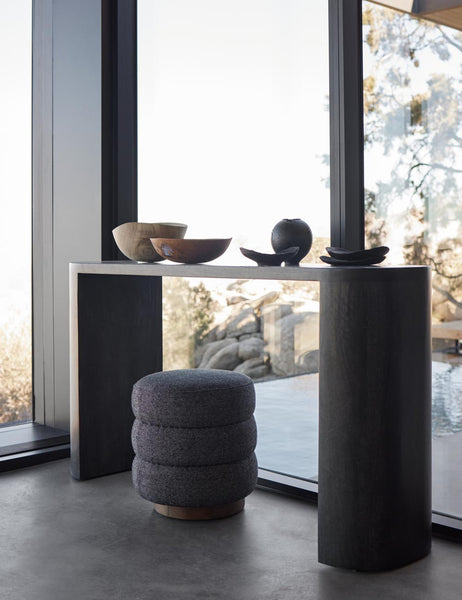 #color::black | The Luna black mango wood oval console table sits in front of floor to ceiling windows with hand-carved wooden bowls sitting atop it and a gray fabric stool underneath.
