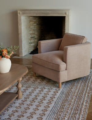 The Belmont apricot linen accent chair sits atop a gray and neutral-toned rug in front of a wooden framed fireplace