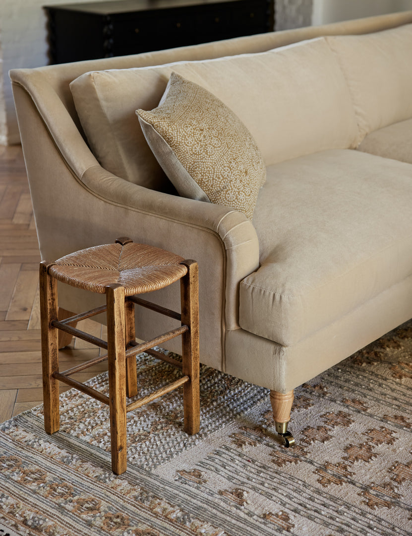 #size::72-W #size:84-W #color::brie-velvet #size::96-W | The Rivington brie beige velvet sofa sits atop a patterned rug next to a stool with a woven base