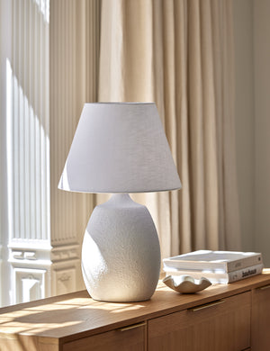 The Pratt white table lamp sits on a natural wooden sideboard next to a stack of books and a white ruffle bowl