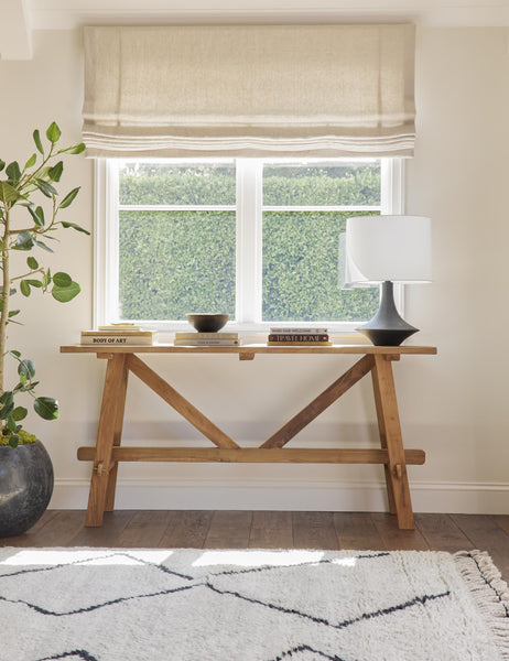 | The Arlene craftsman-style antiqued teak wood console table sits under a window with stacks of books, a black lamp with white shade on top of it, and a white and black Moroccan shag rug below it.
