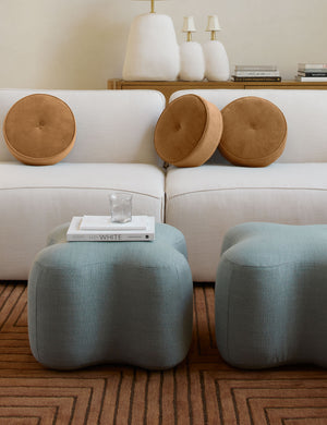 Three Velvet Disc amber Pillows by Sarah Sherman Samuel sit on a white linen sofa in a room with two blue linen ottomans