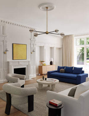The Adeline black and brass retro chandelier sits in a colorful living room with a navy velvet couch, white boucle accent chairs, and a white circular coffee table with accented legs