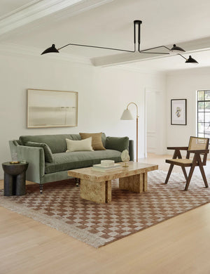 The Irregular ochre checkerboard rug by Sarah Sherman Samuel sits in a living room with a sage velvet couch, a burl wood coffee table, and a woven cane accent chair.