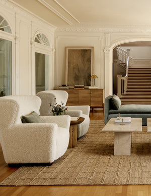 Two Avery Boucle Cream accent chairs sit next to each other atop a textured jute rug with two stone coffee tables atop it