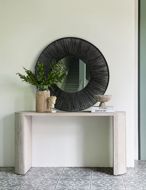 The Luna white-washed oak oval console table sits in an entryway with a black jute mirror, stack of books, and wooden sculptural bowl atop it.