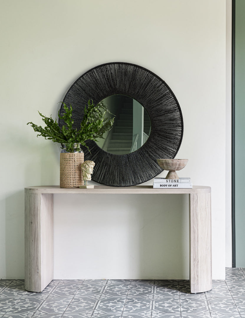 #color::washed-oak | The Luna white-washed oak oval console table sits in an entryway with a black jute mirror, stack of books, and wooden sculptural bowl atop it.