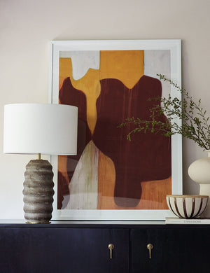 The Ola earth-toned ridged Ceramic Table Lamp by Regina Andrew sits atop a black wooden side table with a warm-toned abstract wall art