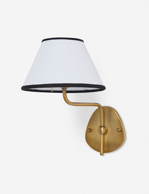 Magdalene brass single sconce with a white linen shade that has a black trim, a round wall mount, and an arched arm