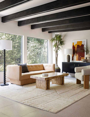 The Fishbone black ebony Floor Lamp by Regina Andrew with a gradated wooden base and linen drum shade sits in a living room with a beige leather couch, a burl wooden coffee table, and a large cream plush rug