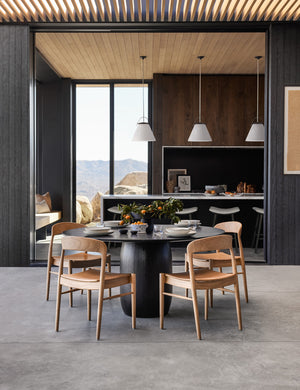 The Ida natural teak wood dining chair sits in an open dining room surrounding a black wood circular dining table with black wood paneling on the walls and wooden beam accents on the ceiling.