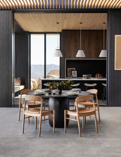#color::Natural | The Ida natural teak wood dining chair sits in an open dining room surrounding a black wood circular dining table with black wood paneling on the walls and wooden beam accents on the ceiling.