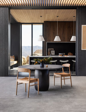 The Maroko black round dining table sits next to two natural wooden dining chairs on a concrete floor under