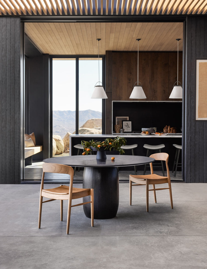 | The Maroko black round dining table sits next to two natural wooden dining chairs on a concrete floor under