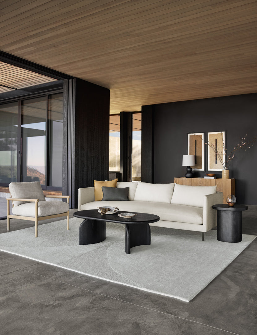 #color::natural #size::72-W #size::84-W #size::96-W | Natural Alaya Sofa sits in a living room with black walls, a black oval coffee table, a textured cream rug, and wooden ceilings