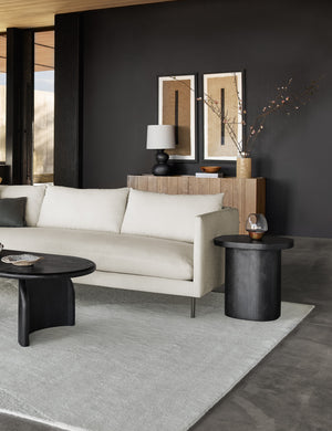 The Luna black wood round side table sits atop an ivory rug next to a natural linen sofa in a room with black walls