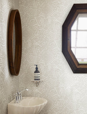 The Malou gray geometric Wallpaper by Malene Barnett is in a bathroom with an octagonal window, a round wooden framed mirror, and a shell-shaped sink