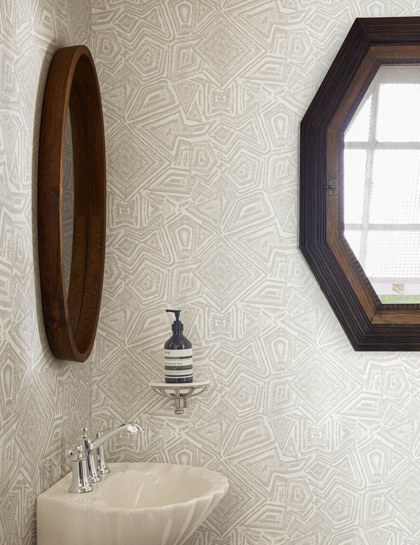 | The Malou gray geometric Wallpaper by Malene Barnett is in a bathroom with an octagonal window, a round wooden framed mirror, and a shell-shaped sink