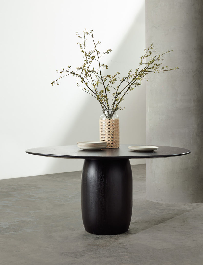 | The Maroko black round dining table sits in a studio with a glass vase and a stack of plates