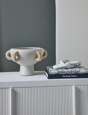 The Clyde terracotta centerpiece with jute handles by Arteriors sits atop a ribbed white sideboard to the left of a stack of books