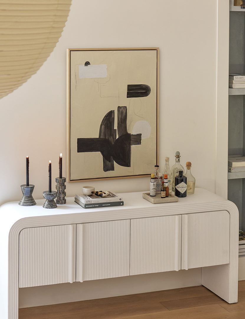 | The In Pursuit Print hangs on a white wall above a white sideboard with stone candle sticks, a bar set, and a stack of books