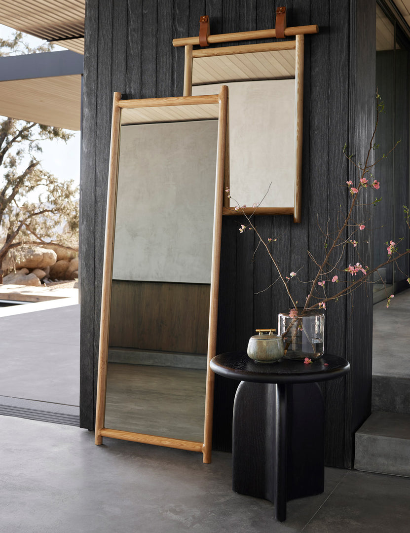 | The kendyl full length mirror lays against a black wood paneled wall next to a wall mirror and a black round side table