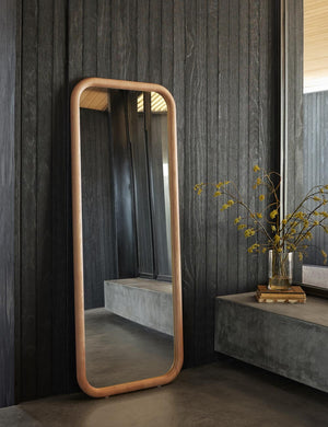 The Bourdon Full Length natural Mirror sits on the floor against a black paneled wall next to floor to ceiling windows
