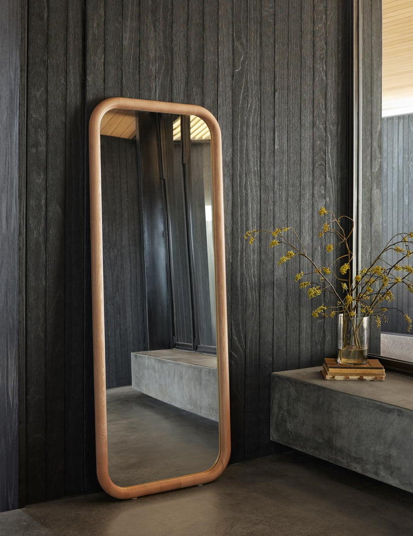 #color::natural | The Bourdon Full Length natural Mirror sits on the floor against a black paneled wall next to floor to ceiling windows