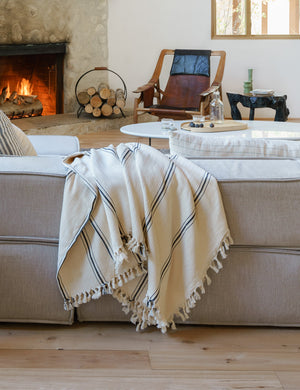 The Monte viso natural and black striped bed cover with fringed ends by House No. 23 sits on a gray linen couch in a rustic living room with a lit fireplace and white stone coffee table