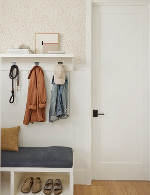 The sommerville goldenrod wallpaper is in an entry room with a hanging coat rack and a white bench with blue cushioning