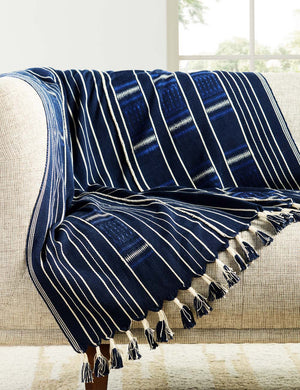 The ajano throw blanket lays atop a natural linen sofa in a room with white walls and large white-framed windows