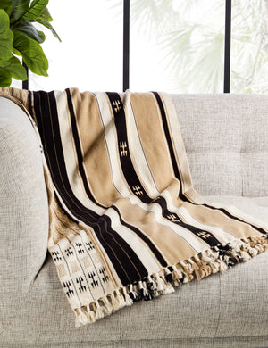 The Sita throw blanket lays on a natural-toned linen sofa in a room with floor to ceiling windows and house plants