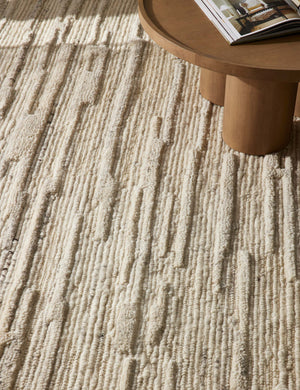 The Rizzoli natural rug sits on a chevron wooden floor under a round wooden coffee table with cylindrical legs