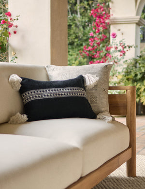 Marchesa black indoor and outdoor lumbar pillow with tasseled corners sits on a natural linen sofa in an outdoor space with pink and green plants in the background