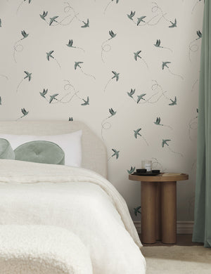 The Sparrow blue and white wallpaper by Rylee and Cru is in a bedroom with a wooden bedside table, a natural upholstered bed with blue throw pillows and curtains as accents.
