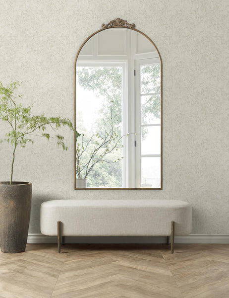 #color::gold | The Tulca gold curved standing mirror with flat bottom edge and traditional scroll detailing is hung on a floral patterned wall above a beige cushioned bench to the right of a stone vase.