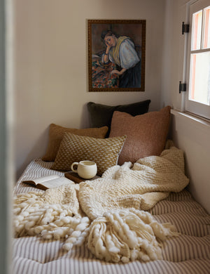 The Olema ivory handwoven throw with fringed ends lays in the corner of a room atop a striped bench with various throw pillows and a portrait painting behind it